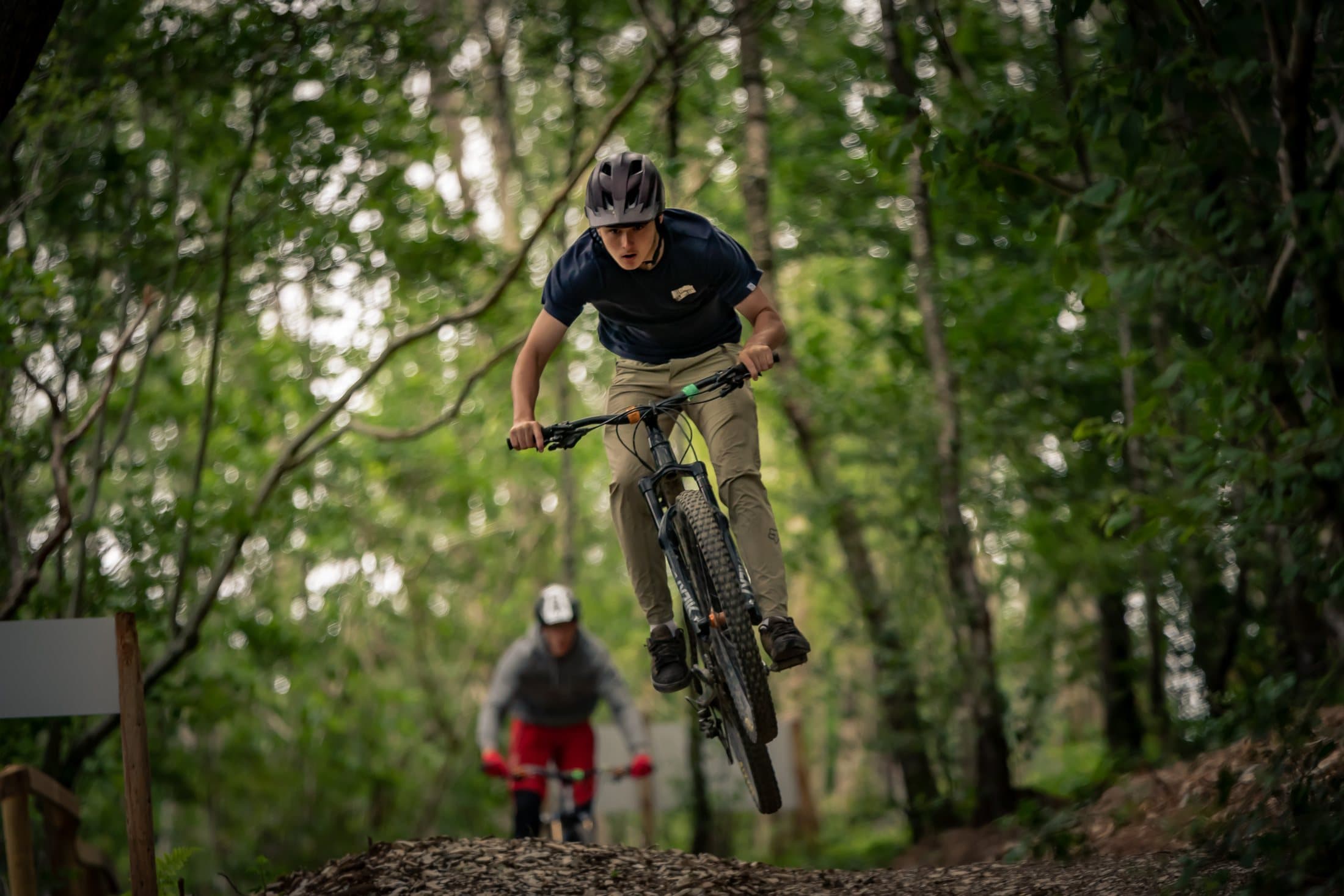 A mountain biker mid-air off a jump, framed by woodland at Hafod Trails